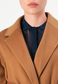 Donna che indossa un cappotto di lana beige sopra una maglia a bottoni blu navy, labbra chiuse, su uno sfondo bianco semplice.