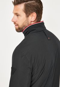 Schwarze Jacke mit hohem Kragen, ausgestattet mit einem rot-weißen Akzentband, strukturiertem Stoff und einem Logo-Patch am linken Ärmel.