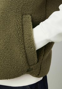 Gilet en polaire de couleur vert olive avec un extérieur à texture douce et duveteuse. Il dispose de poches latérales et d'une petite étiquette de marque. Superposé sur une chemise blanche.