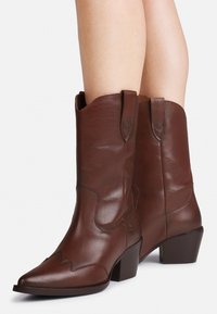 Botine din piele de cowboy maro, cu lungimea până la gambe, vârfuri ascuțite, cusături decorative și tocuri pătrate purtate pe picioare goale, pe un fundal simplu.
