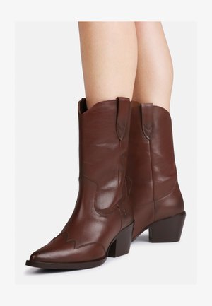 Botas de vaquero de cuero marrón hasta la mitad de la pantorrilla con puntas afiladas, costuras decorativas y tacones cuadrados, llevadas sobre piernas desnudas contra un fondo sencillo.