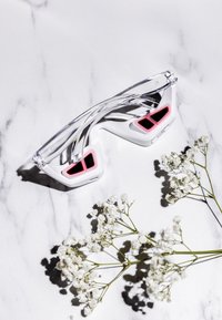 Okulary przeciwsłoneczne z przezroczystą oprawką, czarnymi soczewkami obramowanymi różowymi akcentami, zakrzywionymi zausznikami, umieszczone na marmurowej powierzchni z białymi kwiatami.