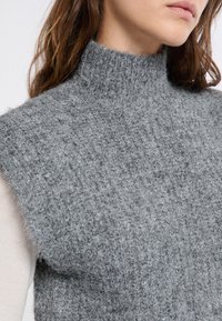 Maglione grigio lavorato a maglia senza maniche con collo alto. La texture è morbida e pelosa, con una vestibilità ampia e rilassata.