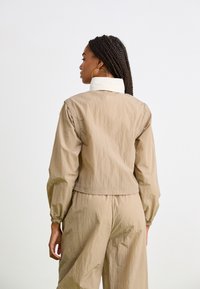 Beige croppet jakke med hvit krage, med elastiske mansjetter og teksturert stoff, båret over matchende beige bukser.