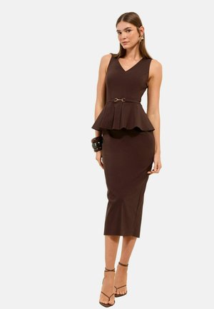 Frau trägt ein ärmelloses braunes Peplum-Kleid mit Midirock, schwarzen Armreifen und Riemchen-Absätzen vor einem schlichten hellen Hintergrund.