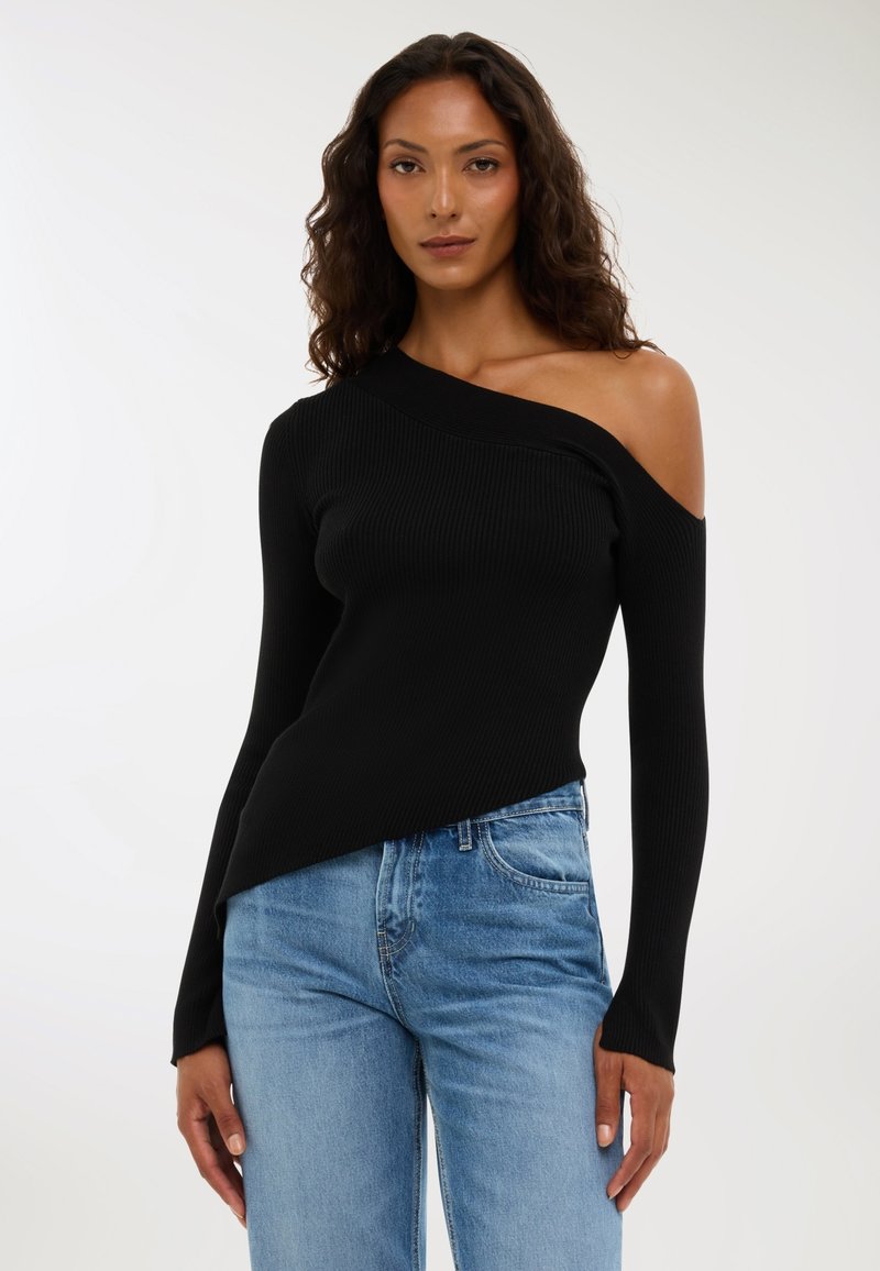 Kobieta z kręconymi włosami w czarnym swetrze z jednym odkrytym ramieniem i jasnoniebieskich dżinsach, stojąca na jednolitym tle.