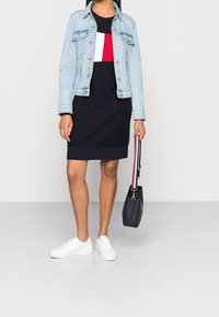 Jeansjacke über einem marineblauen Kleid mit einem rot-weißen Blockdesign; kombiniert mit weißen Sneakers und einer kleinen dunklen Tasche. Glatte Texturen, lässiger Stil.