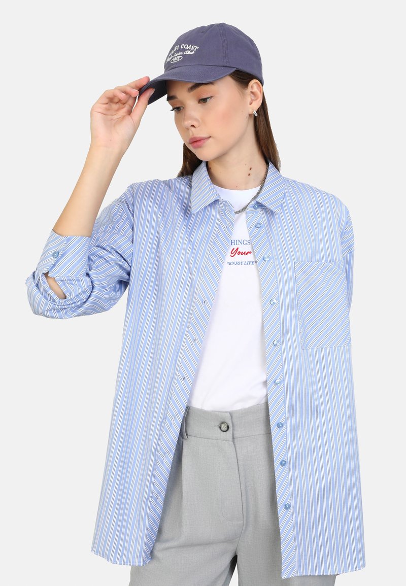 Blaut gestreiftes Hemd in Weiß mit Tasche, über einem weißen T-Shirt getragen. Graue Hose. Modell trägt eine marineblaue Kappe mit Text.