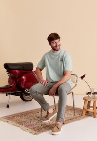 Lichtgroene t-shirt met korte mouwen met een palmboomafbeelding. Grijze slim-fit broek en beige sneakers. Vintage rode scooter op de achtergrond.