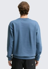 Niebieska bluza z okrągłym dekoltem, długimi rękawami i ściągaczami. Gładka faktura z luźnym krojem, bez widocznych wzorów ani logo.