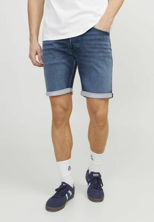 Hombre vestido con pantalones cortos de mezclilla azul oscuro arremangados, calcetines blancos hasta el tobillo con un logo azul, y zapatillas deportivas azul marino con Rayas azul claro.