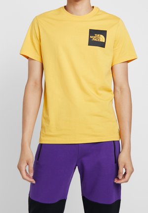 Osoba ubrana w żółtą koszulkę The North Face i fioletowe spodnie z czarnymi łatkami na kolanach, stojąca na jednolitym tle.
