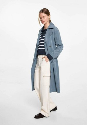 Trench coat bleu deschis cu un design cu două rânduri de nasturi, purtat peste un top bleu închis cu dungi albe și pantaloni cargo crem. Textură netedă.