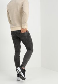 Jeans negros de corte slim con una textura desgastada, combinados con un suéter beige y zapatillas negras con suelas blancas, vistos desde atrás.