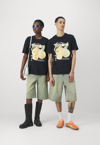 T-shirts graphiques noirs avec l'impression "Florida Tropical", shorts en denim vert clair, bottes noires hautes jusqu'aux genoux et baskets oranges avec rayures.