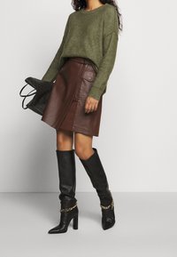 Pull en tricot vert olive, jupe asymétrique en cuir marron, bottes hautes en cuir noir avec un accent chaîne. Sac fourre-tout noir à motif.