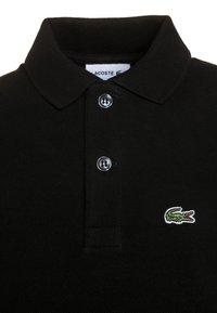 Polo shirt nera con tessuto a maglia testurizzato, due bottoni argentati e logo di coccodrillo ricamato in verde sul lato sinistro.