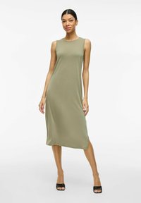 Beżowa sukienka midi bez rękawów uszyta z miękkiego materiału, z okrągłym dekoltem i bocznymi rozporkami, zestawiona z czarnymi sandałami na obcasie.