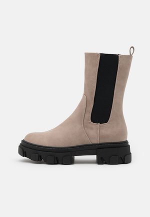 Botte beige mi-mollet avec semelle crantée noire épaisse, panneau latéral élastique et languette à l'arrière.