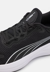 Zapatilla deportiva negra con parte superior de malla, cordones negros gruesos y rayas blancas. Presenta un diseño texturizado y una suela de goma con amortiguación ligera.