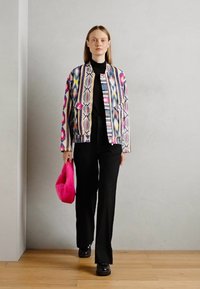 Vrouw die binnenshuis loopt in een multicolor geblokte jas, zwarte coltrui, zwarte broek, zwarte schoenen en een felroze handtas vasthoudt.