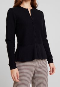 Giacca nera con zip realizzata in tessuto testurizzato, con orlo a peplum, dotata di scollatura rotonda e maniche lunghe, indossata sopra pantaloni a quadri.
