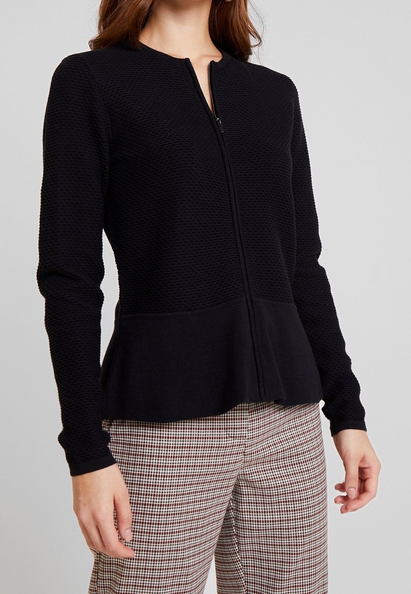 Giacca nera con zip realizzata in tessuto testurizzato, con orlo a peplum, dotata di scollatura rotonda e maniche lunghe, indossata sopra pantaloni a quadri.