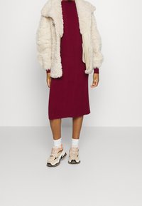 Veste en fausse fourrure crème surdimensionnée, robe en maille côtelée bordeaux, chaussettes blanches et baskets beige avec des accents orange.