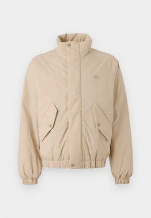 Beige padded jacket with a high collar, elastic cuffs, and two angled front pockets. Features brand logo stitched in matching color.