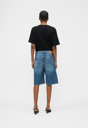 Osoba z zaplecionymi włosami, ubrana w czarny T-shirt, długie krótkie spodenki jeansowe i czarne sandały na obcasie, stoi tyłem na jednolitym tle.