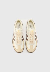 Des chaussures de sport blanches et beiges avec trois rayures marron, un bout arrondi, une semelle texturée et des lacets de couleurs coordonnées.