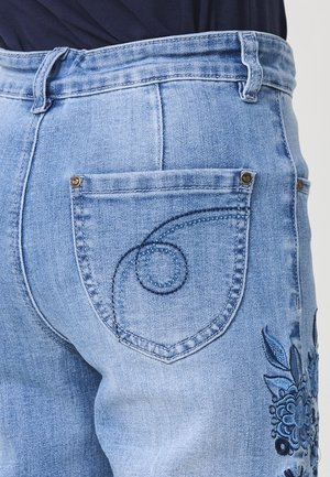 Helle blaue Jeans mit einem bestickten Blumenmuster an der Seite und einem genähten Wirbelmuster auf der Gesäßtasche.