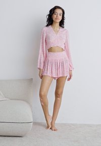 Top cropped com padrão rosa e mangas longas, combinado com uma saia mini franzida a condizer. Modelo está descalça sobre uma superfície neutra com carpete.
