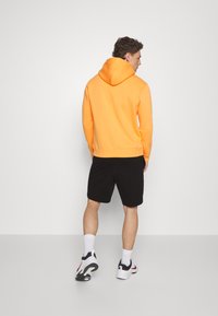 Sudadera naranja con bolsillo tipo canguro, pantalones cortos negros y zapatillas blancas. La sudadera cuenta con capucha ajustable y puños acanalados.