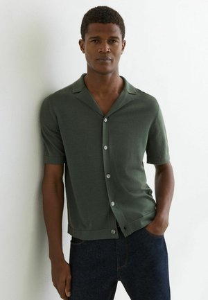 Mężczyzna w ciemnozielonej koszuli na krótki rękaw z guzikami oraz ciemnych dżinsach stoi oparty o białą ścianę, z jedną ręką w kieszeni.