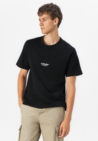 Sort bomulds T-shirt med korteærmer og rund hals. Har hvidt trykt logo på brystet. Båret med lysebeige cargo bukser.