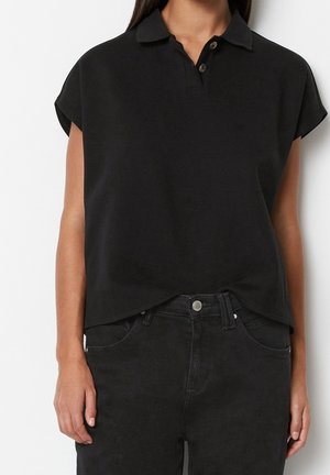 Vrouw met een zwart poloshirt met korte mouwen en zwarte jeans, staand tegen een effen witte achtergrond, zichtbaar van schouders tot heupen.