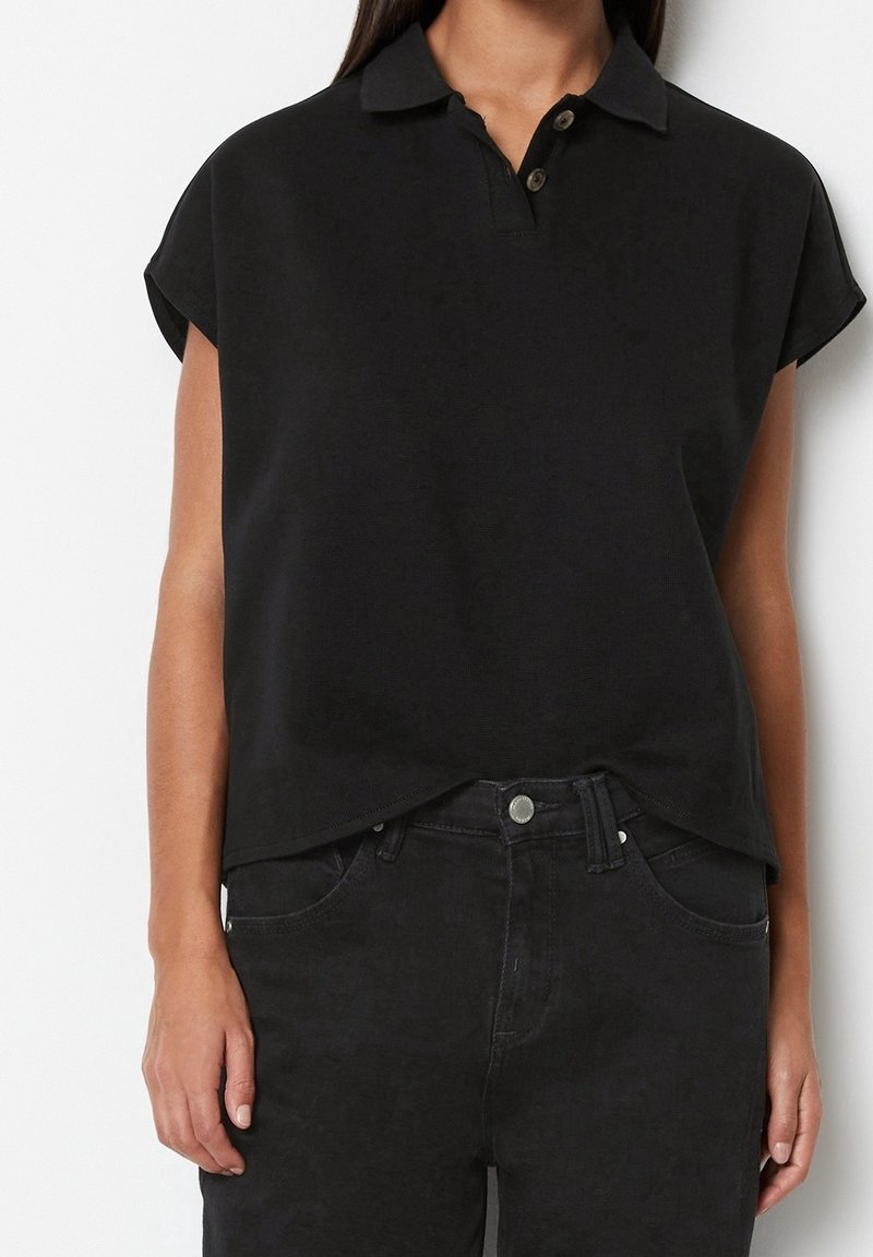 Femme portant un polo noir à manches courtes et un jean noir, debout devant un fond blanc uni, vue des épaules aux hanches.