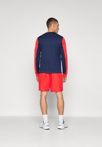 Jūras spēku pārslēgi ar sarkaniem piedurknēm un atbilstošām sarkanām šortiem. Apģērbs raksturo vienkāršu dizainu un gludu auduma faktūru. Balti sporta apavi papildina izskatu.