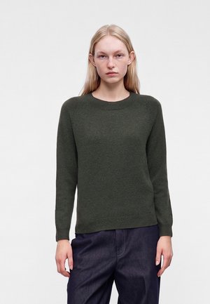 Femme aux longs cheveux blonds portant un pull en laine vert foncé et un pantalon sombre sur un fond gris clair uni.