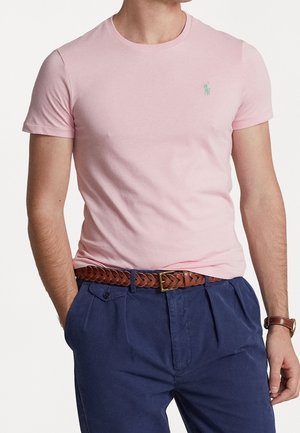 Mann trägt hellrosa T-Shirt mit Rundhalsausschnitt, dunkelblaue Hose mit braunem geflochtenem Gürtel und braune Armbanduhr, dargestellt von Hals bis Taille.