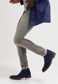 Stivali alla caviglia blu scamosciati con suole in gomma marrone, abbinati a pantaloni slim-fit grigio chiaro. Le calzature presentano lacci e una finitura testurizzata.