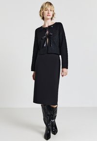 Donna che indossa un cardigan nero a trama con lacci, una gonna nera aderente midi e stivali neri lucidi al ginocchio, in piedi su uno sfondo semplice.