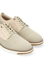 Chaussures beiges en dentelle perforée à lacets avec un accent en cuir lisse, semelle blanche et détails marron au niveau des orteils et du talon.
