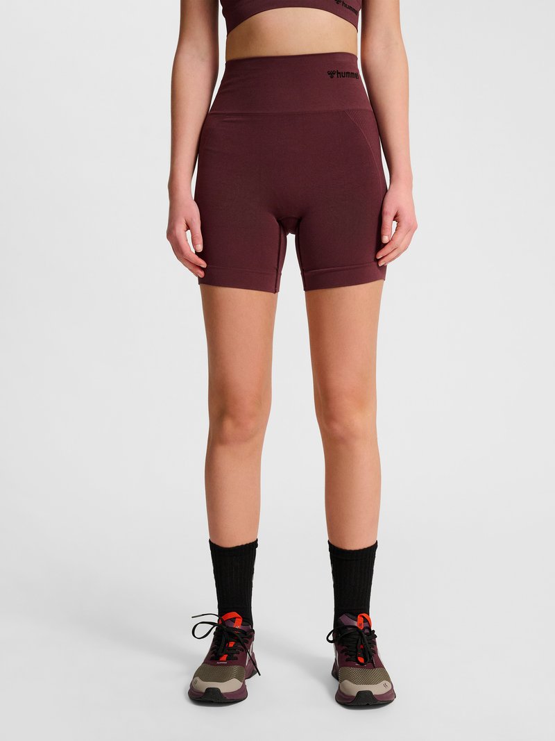 Person trägt weinrote, hoch geschnittene nahtlose Biker-Shorts, schwarze Socken und mehrfarbige Sportschuhe, steht vor einem schlichten Hintergrund.
