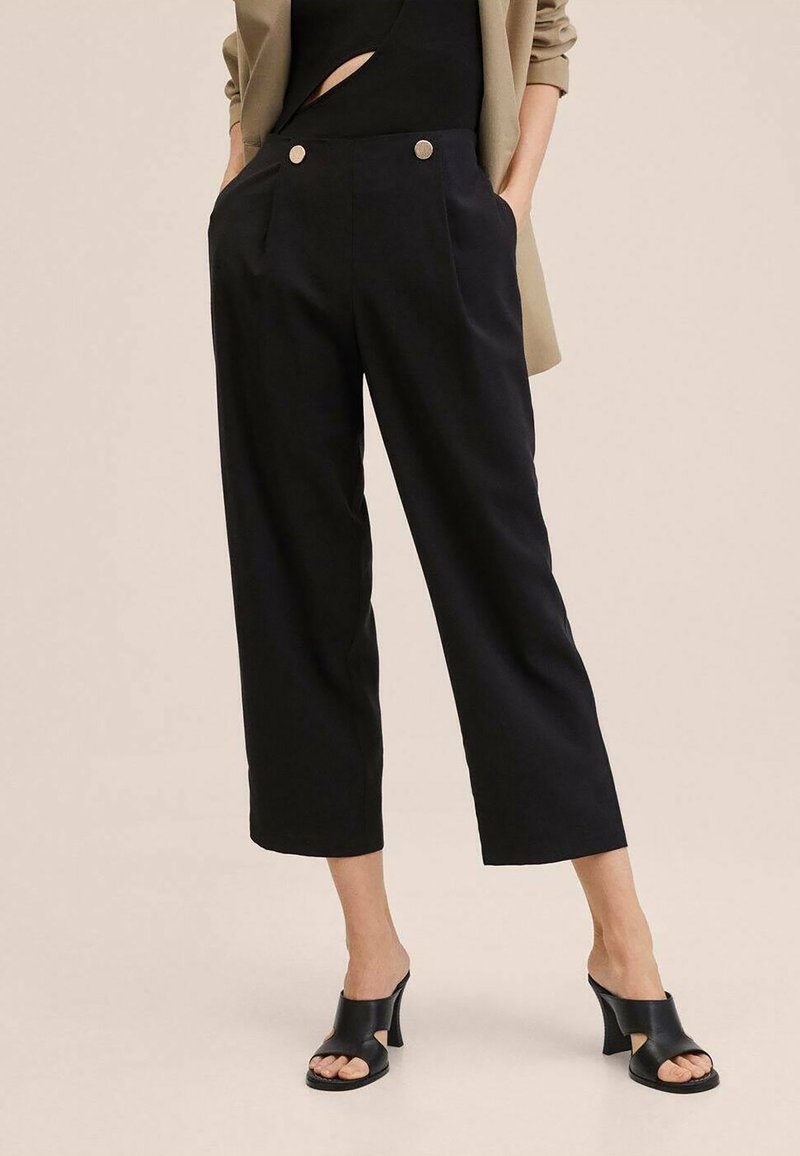 Femme portant un pantalon noir court, taille haute, à jambes larges avec des boutons, et des mules noires à talons ouvertes à l'avant, sur un fond uni.