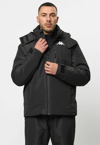 Schwarze wasserdichte Jacke mit hohem Kragen, Reißverschluss und Klettverschlüssen. Mit einem Logo auf der Brust und verstellbarem Kapuzenhood.