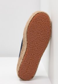 Podeszwa buta espadryli z teksturowaną, brązową gumową podeszwą i plecioną lamówką z juty, ustawiona pod kątem na białej powierzchni.