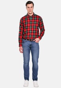 Czerwona i zielona koszula w kratę z guzikami, dopasowane niebieskie dżinsy i białe trampki. Koszula ma kołnierzyk i guziki z przodu.