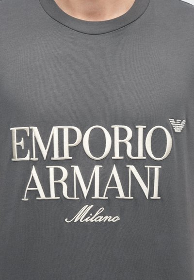 Sivé bavlnené tričko s veľkým bielym vyšívaným nápisom „EMPORIO ARMANI“ a menším nápisom „Milano“ v kurzíve, akcentové logo na ramene.
